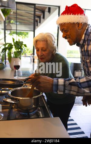 Heureux couple d'amis divers dans le chapeau de santa préparant la nourriture dans la cuisine ensoleillée à la maison Banque D'Images