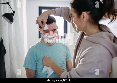 Mère appliquant de la mousse à raser sur le visage de son fils. Jeune homme trisomique apprenant à se raser. Banque D'Images