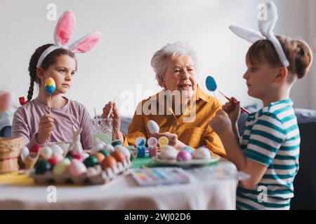 Grand-mère avec de petits enfants décorant des œufs de pâques à la maison. Tradition de peindre les oeufs avec pinceau et colorant d'oeuf de pâques. Banque D'Images