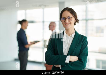 Jeune femme chef de la direction réussie dans les lunettes et les vêtements de forme regardant la caméra avec le sourire contre deux collègues Banque D'Images
