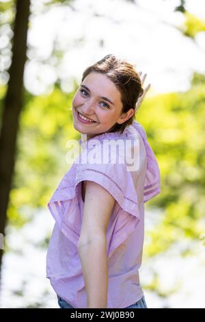 Cette image montre une jeune femme qui se tourne pour sourire par-dessus son épaule, ses yeux brillants de joie et de jeu. Elle est dehors par une journée ensoleillée, avec des arbres fournissant un fond de lumière tapissée. Son haut violet clair ajoute une touche de couleur et une sensation légère et aérée à l'image, suggérant une journée joyeuse et détendue. Jeune femme ludique souriant sur l'épaule. Photo de haute qualité Banque D'Images