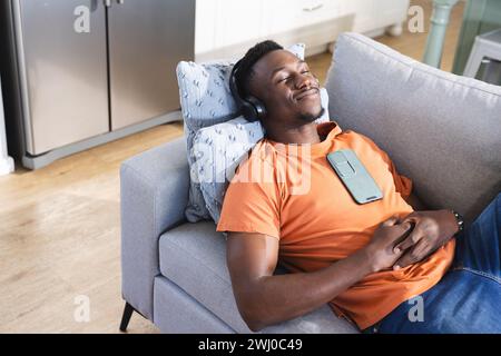 Homme afro-américain souriant sur le canapé dans les écouteurs, écouter de la musique sur smartphone, copier l'espace Banque D'Images