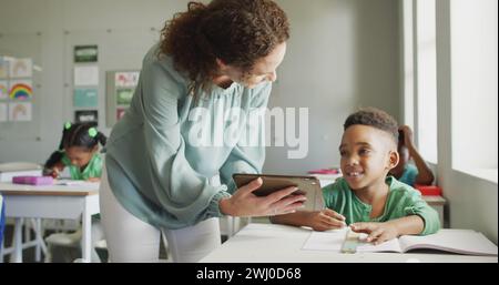 Image du traitement des données sur une enseignante et un écolier heureux et diversifié utilisant une tablette en classe Banque D'Images