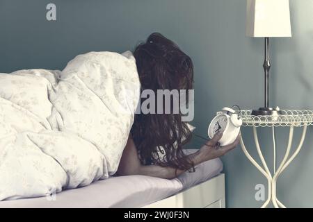 Femme se réveille sur le lit en regardant le réveil. Mettre le réveil dans la chambre à la maison Banque D'Images