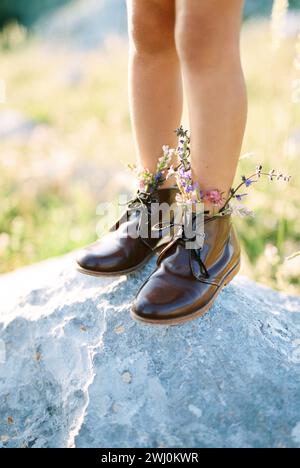 Les pieds d'une petite fille en bottes avec des fleurs sauvages se tiennent sur une pierre. Rogné Banque D'Images