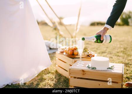 Le marié verse le champagne dans des verres sur une boîte en bois avec un gâteau à côté de la mariée. Rogné Banque D'Images