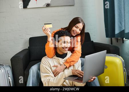 heureux couple multiethnique magasinant en ligne, tenant une carte de crédit, avec un ordinateur portable près des bagages Banque D'Images