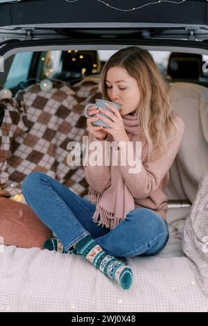 Fille assise les jambes croisées sur une couverture dans le coffre d'une voiture et boit du café d'une tasse Banque D'Images