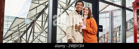 Jeune couple diversifié souriant tout en tenant un café pour aller et entrer dans une auberge moderne, bannière Banque D'Images