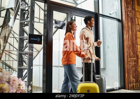 heureux couple diversifié avec des bagages de voyage souriant et entrant dans une auberge moderne, tenant le café à emporter Banque D'Images