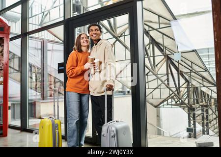 Jeune couple diversifié avec des bagages de voyage souriant et entrant dans une auberge moderne, tenant le café à emporter Banque D'Images