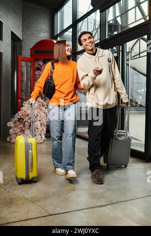 Jeune couple diversifié entrant heureux dans un bâtiment moderne avec leurs valises roulantes, auberge Banque D'Images