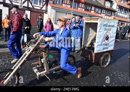 Herbstein, Allemagne. 12 février 2024. Une construction de cyclomoteur en bois tire une remorque au Herbstein Springerzug, qui recommande « d'économiser l'électricité en conduisant un moteur à combustion ». Les tailleurs de pierre tyroliens ont apporté la tradition à Herbstein au XVIIe siècle. Plus de 50 personnes ont participé au traditionnel défilé du lundi Rose dans la ville du district de Vogelsberg. Crédit : Uwe Zucchi/dpa/Alamy Live News Banque D'Images