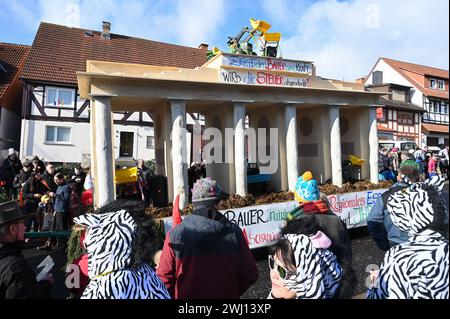 Herbstein, Allemagne. 12 février 2024. Un flotteur à motif avec des tracteurs sur la réplique de la porte de Brandebourg rappelle les récentes manifestations des agriculteurs lors du défilé Herbstein Springer. Les tailleurs de pierre tyroliens ont apporté la tradition à Herbstein au XVIIe siècle. Plus de 50 chars ont pris part au traditionnel défilé Rose Monday dans la ville du district de Vogelsberg. Crédit : Uwe Zucchi/dpa/Alamy Live News Banque D'Images