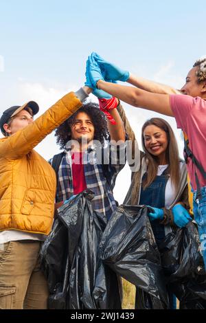 L'image montre un groupe de bénévoles enthousiastes célébrant leur nettoyage environnemental réussi. L'équipe, composée d'un mélange diversifié d'individus, est vue lever les mains gantées ensemble dans un geste d'unité et d'accomplissement. Ils tiennent des sacs poubelles noirs, indiquant la litière qu'ils ont ramassée. Le groupe comprend un homme caucasien dans une veste jaune, un homme afro-américain aux cheveux bouclés dans une chemise à carreaux, une femme asiatique dans une salopette en denim et une personne aux cheveux rosâtres dans un haut rouge. Leurs visages sont remplis de sourires, reflétant la fierté de leur impact positif sur l'environnement. VO Banque D'Images