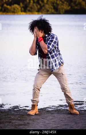 Cette image représente un homme afro-américain dans un moment de détresse ou de pensée profonde. Il se tient près d'un front de mer calme, couvrant son visage avec ses mains, ce qui peut suggérer tristesse, déception ou fatigue. La tenue de l'homme comprend une chemise à carreaux sur un t-shirt Bordeaux, un pantalon kaki et des bottes brunes robustes, suggérant un cadre décontracté et extérieur. Ses cheveux noirs bouclés sont proéminents, et il porte un bracelet rouge, qui se distingue comme une tache de couleur vive par rapport à sa tenue autrement terreuse. L'image transmet un sentiment de solitude et d'émotion, dans le cadre tranquille d'un plan d'eau Banque D'Images