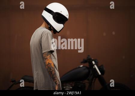Personne tatouée avec casque face à une moto noire personnalisée contre un mur marron. Banque D'Images