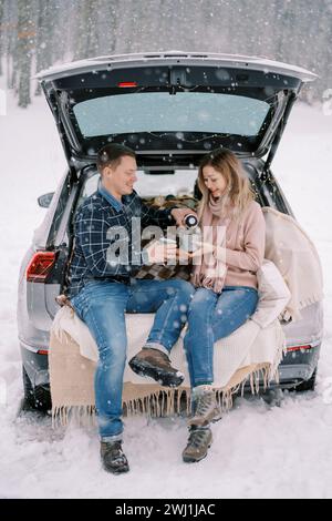 Mari verse le café d'un thermos dans sa tasse de femme tout en étant assis avec elle dans le coffre d'une voiture sous une chute de neige dans les Fores Banque D'Images