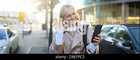 Souriant jeune femme moderne, marchant dans la rue, chats vidéo avec un ami, dit bonjour, la main des vagues à la caméra du téléphone portable, parlant t Banque D'Images