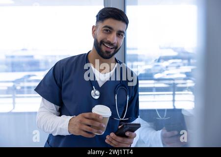 Portrait de médecin homme biracial heureux utilisant smartphone et tenant une tasse de café dans le bureau de l'hôpital Banque D'Images