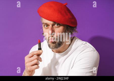 Drôle homme d'âge moyen portant béret rouge tient le rouge à lèvres dans sa main sur fond violet Banque D'Images