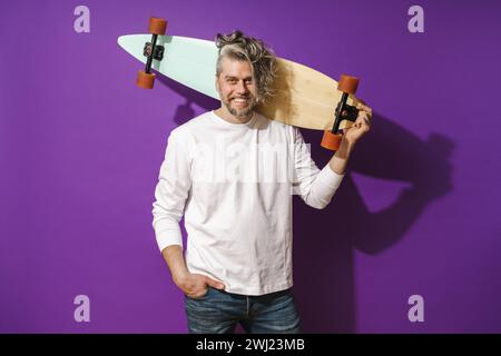 Joyeux homme d'âge moyen avec longboard portant un sweat-shirt blanc sur fond violet Banque D'Images