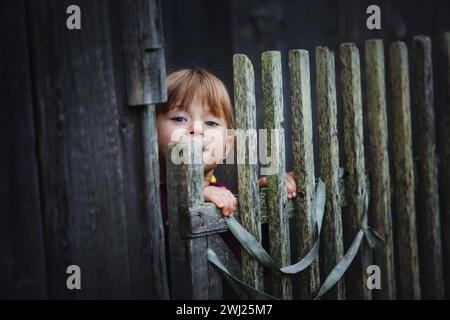 Une petite fille mignonne jette un coup d'œil par derrière une clôture en bois rustique. Banque D'Images