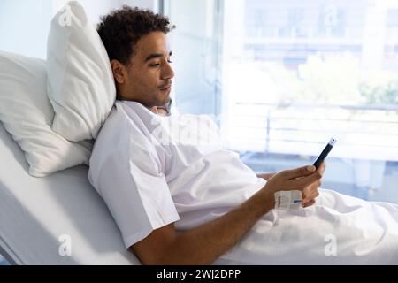 Patient masculin biracial utilisant smartphone avec perfusion intraveineuse dans le lit dans la chambre d'hôpital ensoleillée Banque D'Images