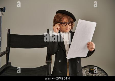 garçon élégant dans des lunettes et béret lisant scénario sur papier sur gris, enfant en tant que réalisateur de cinéaste Banque D'Images