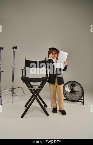 garçon élégant dans des lunettes et béret lisant scénario sur papier sur gris, enfant en tant que réalisateur de cinéaste Banque D'Images