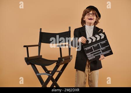 garçon cinéaste excité dans des vêtements à la mode pose joyeusement avec clapet dans la main contre le mur beige Banque D'Images