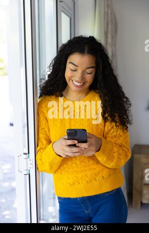 Heureuse femme biraciale en pull jaune à l'aide d'un smartphone à la maison Banque D'Images