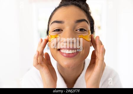 Portrait de femme biraciale heureuse en peignoir appliquant le masque pour les yeux dans la salle de bain ensoleillée Banque D'Images