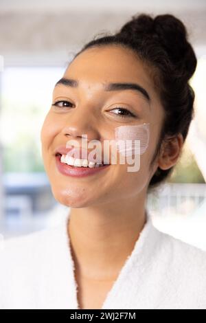 Portrait de femme biraciale heureuse en peignoir appliquant le masque facial dans la salle de bain ensoleillée Banque D'Images