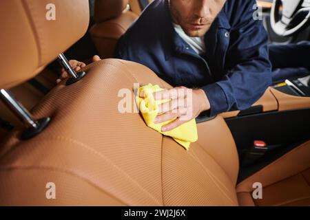 vue rognée de l'ouvrier dévoué en uniforme bleu confortable nettoyant attentivement la voiture avec un chiffon jaune Banque D'Images
