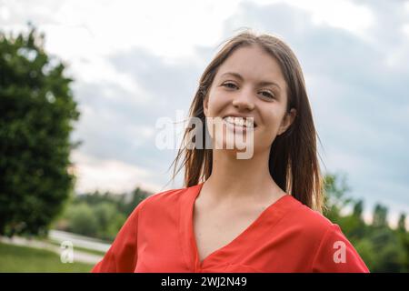 Une jeune femme joyeuse dans une robe rouge vibrante profite d'une journée ensoleillée dans le parc, exsudant le bonheur et la confiance. Banque D'Images