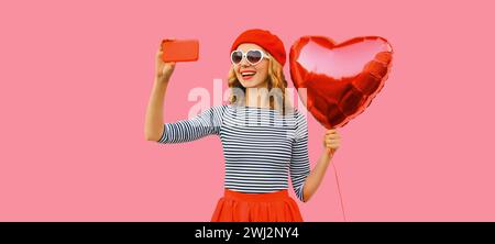 heureuse jeune femme souriante prenant selfie avec un téléphone portable tenant un ballon en forme de coeur rouge portant un béret français, lunettes de soleil sur fond de studio rose Banque D'Images