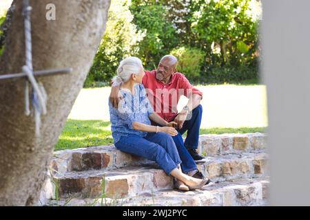 Heureux couple senior diversifié embrassant et assis sur les escaliers dans le jardin ensoleillé Banque D'Images