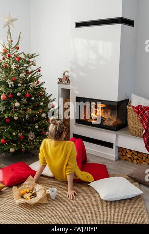 Femme heureuse est assise à côté d'une cheminée incandescente dans un salon confortable, orné d'un arbre de Noël Banque D'Images