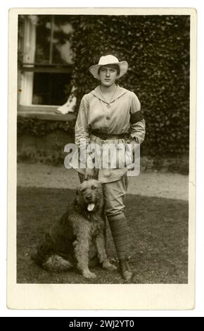 Carte postale originale de l'époque de la première Guerre mondiale d'une landgirl appelée Esme Greene portant une tunique beige indiquant les tâches agricoles générales (par opposition à l'uniforme blanc pour la laiterie) et un brassard de service de 3 mois. Elle porte des culottes, une tunique au genou (avec une ceinture). Elle est debout devant une maison de campagne ou une ferme, avec un chien appelé Kim, daté de 1917, Easton à Gordano, près de Bristol, Royaume-Uni Banque D'Images