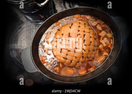 Rôti de porc avec écorce en forme de losange et lardé avec des clous de girofle dans une cocotte avec des légumes et du bouillon sur une cuisinière noire, COO Banque D'Images