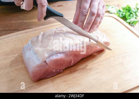 La croûte d'un rôti de porc cru est incisée en forme de diamant avec un couteau tranchant sur un plan de cuisine en bois, préparation pour la cuisson A. Banque D'Images