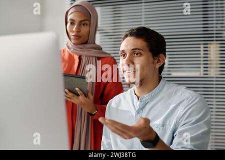 Jeune analyste musulman confiant expliquant les données sur l'écran d'ordinateur à une collègue en hijab lors de la présentation des points du projet Banque D'Images