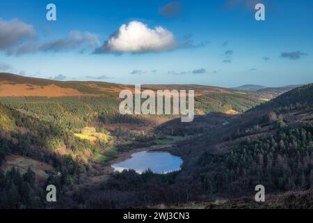 Lac Glendalough et cimetière dans la vallée. Forêt sur les collines illuminées par la lumière du soleil. Randonnée dans les belles montagnes de Wicklow d'automne, Irlande Banque D'Images