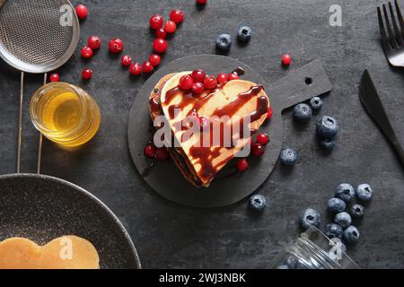 Planche d'ardoise avec de savoureuses crêpes en forme de coeur, de baies et de confiture sur fond sombre. Célébration de la Saint-Valentin Banque D'Images