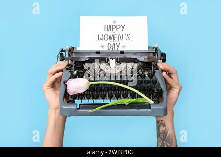 Mains tenant machine à écrire vintage, tulipe et carte postale festive avec texte HAPPY WOMEN'S DAY sur fond bleu Banque D'Images