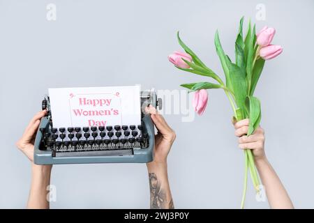 Mains tenant une machine à écrire vintage, des tulipes et une carte postale festive avec texte HAPPY WOMEN'S DAY sur fond gris Banque D'Images