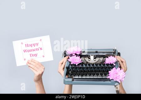 Mains tenant une machine à écrire vintage, des fleurs et une carte postale festive avec texte HAPPY WOMEN'S DAY sur fond gris Banque D'Images