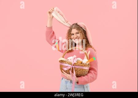 Belle jeune femme heureuse dans les oreilles de lapin avec panier de Pâques sur fond rose Banque D'Images