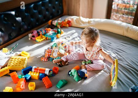 Petite fille assise avec un xylophone et une baguette dans ses mains sur le lit entourée de peluches Banque D'Images